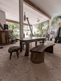A rustic, open-air dining area with a wooden table and seating set on a sandy floor. The space is surrounded by lush greenery and palm trees, visible through the large openings. The ceiling is supported by white columns with wooden beams and features a ceiling fan for ventilation.