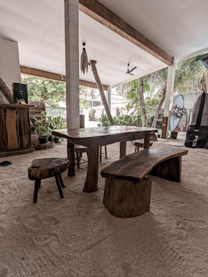 A rustic, open-air dining area with a wooden table and seating set on a sandy floor. The space is surrounded by lush greenery and palm trees, visible through the large openings. The ceiling is supported by white columns with wooden beams and features a ceiling fan for ventilation.