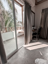 A room with a glass sliding door leading to a balcony with a view of palm trees. Inside, there is a small desk with a chair and a mirror on the wall. The floor is made of polished concrete, and long curtains hang beside the glass door.