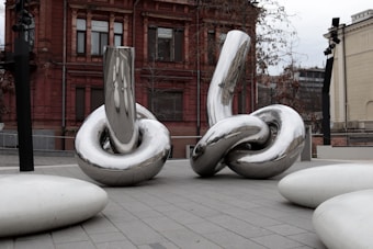 Large, intertwined metallic sculptures with a polished, reflective surface stand in an urban outdoor setting. The background features a red brick building with windows and some bare trees, indicating a city environment. The sculptures have a modern, abstract design and the ground is paved with tiles.