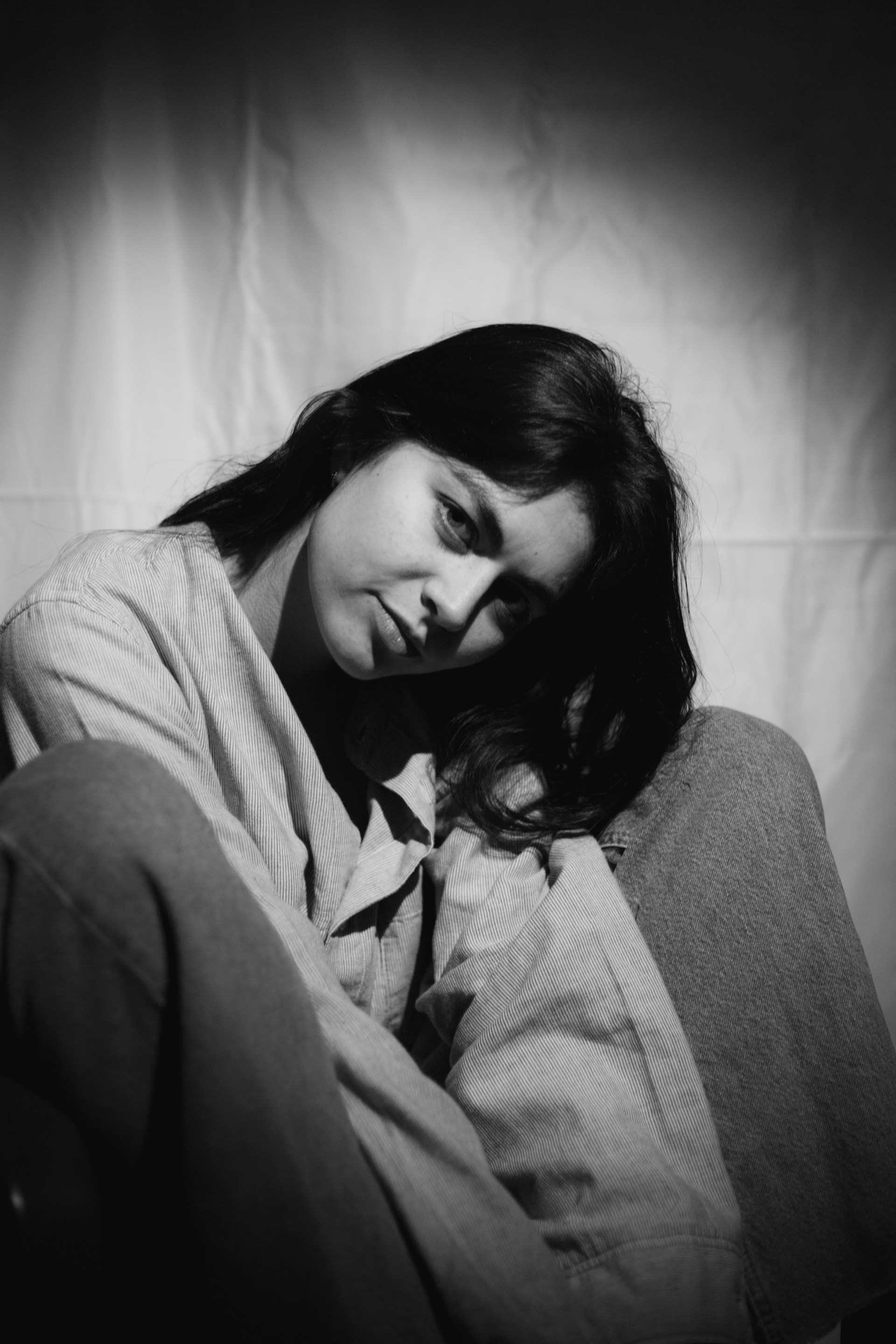 A black and white portrait of a woman with long dark hair, wearing a buttoned shirt and jeans. She is sitting down, slightly leaning forward with her head tilted to the side. The background is plain with soft lighting that creates shadows, giving a moody and introspective feel.
