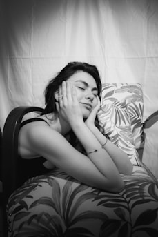A person with long dark hair is leaning against a patterned cushion with their eyes closed and hands resting on their cheeks. The setting appears to be relaxed and calm, with soft lighting adding a serene atmosphere.