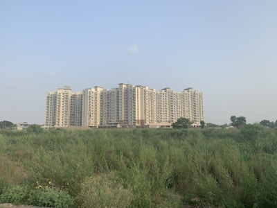 A large residential apartment complex stands tall in the background, consisting of multiple similar-looking high-rise buildings. In the foreground, there is an open field with dense green vegetation and a clear, pale sky overhead.