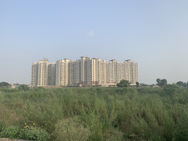 A large residential apartment complex stands tall in the background, consisting of multiple similar-looking high-rise buildings. In the foreground, there is an open field with dense green vegetation and a clear, pale sky overhead.