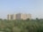 A large residential apartment complex stands tall in the background, consisting of multiple similar-looking high-rise buildings. In the foreground, there is an open field with dense green vegetation and a clear, pale sky overhead.