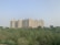 A large residential apartment complex stands tall in the background, consisting of multiple similar-looking high-rise buildings. In the foreground, there is an open field with dense green vegetation and a clear, pale sky overhead.