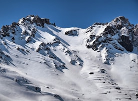 Snow-covered mountainous terrain with jagged peaks and deep shadows. Ski tracks are visible, indicating recent activity in the soft snow. The sky is clear, suggesting bright and favorable weather conditions.