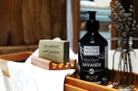 An artisanal soap bar with 'Savons Arthur' engraved on it sits on a wooden soap dish. Next to it is a bottle of liquid soap labeled 'Savons Arthur Bio Lavande.' Both items rest on a wooden tray with a white cloth. A glass bottle with dried lavender adds a rustic touch to the composition.