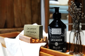 An artisanal soap bar with 'Savons Arthur' engraved on it sits on a wooden soap dish. Next to it is a bottle of liquid soap labeled 'Savons Arthur Bio Lavande.' Both items rest on a wooden tray with a white cloth. A glass bottle with dried lavender adds a rustic touch to the composition.