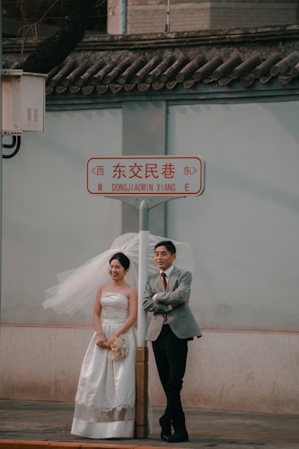 A couple stands under a street sign, with the woman wearing a white wedding dress and holding a bouquet, while the man is dressed in a suit with a red tie. They appear to be posing for a photo with the bride smiling and the groom leaning against a pole. The background features a traditional tiled roof.