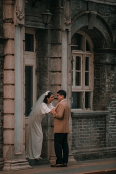 A bride in a white gown and veil leans out of a building's doorway, holding hands with a man in a tan suit. The setting is an old stone building with arched windows and a vintage lamp above the door.
