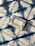 A bar of natural soap in a package lies on a patterned fabric surface. The soap packaging displays text and nature-related symbols, suggesting it is an organic product. The fabric features a bold, abstract blue and white pattern with a textured appearance.