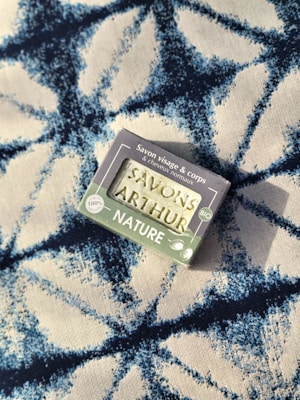 A bar of natural soap in a package lies on a patterned fabric surface. The soap packaging displays text and nature-related symbols, suggesting it is an organic product. The fabric features a bold, abstract blue and white pattern with a textured appearance.