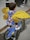 A child rides a blue bicycle with training wheels along a wet sidewalk. The child is wearing a colorful jacket with a patchwork design and large, shiny golden wings on their back, which appear to be inflatable or decorative. The scene is urban, with pavement and a curb in the background.