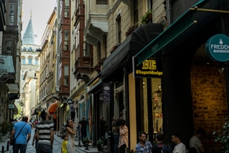 A narrow street is lined with historic buildings featuring ornate facades and a mix of architectural styles. People are walking and standing in groups, engaging in various activities. A prominent tower with a conical roof is visible in the distance, providing a picturesque backdrop. Shop signs and awnings are present along the street, adding to the urban atmosphere.