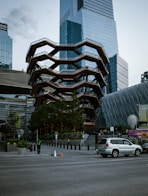 A modern architectural structure with geometric patterns stands prominently in an urban setting. The building features interconnected staircases and platforms, surrounded by high-rise buildings with reflective glass facades. Below, a city street with pedestrians, a silver SUV, and greenery can be observed.