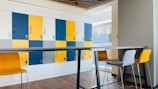 A modern office space featuring a row of colored lockers with a combination of blue, yellow, and white doors. Each locker is marked with a number and a keypad lock. In front, there is a high table with matching high chairs in mustard yellow and gray, set against a wood-paneled floor. Natural light is streaming in from the large windows on the right.