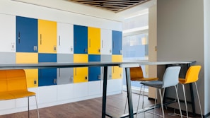 A modern office space featuring a row of colored lockers with a combination of blue, yellow, and white doors. Each locker is marked with a number and a keypad lock. In front, there is a high table with matching high chairs in mustard yellow and gray, set against a wood-paneled floor. Natural light is streaming in from the large windows on the right.