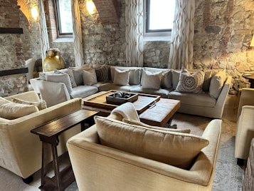A cozy living room features rustic stone walls and arched brick details. The space is furnished with a large sectional sofa and several soft armchairs, all adorned with an assortment of cushions. A wooden coffee table sits centrally, atop a light-colored area rug, and a decorative vase adds a touch of elegance. Soft lighting from wall sconces enhances the warmth of the room.