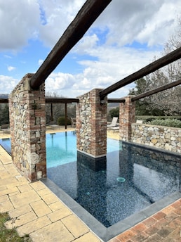 A rectangular swimming pool features two sections with the closer section having darker tiles and a more shallow appearance. The pool is bordered by stone and brick columns supporting wooden beams, creating an open, rustic look. Surrounding the pool is a patio area with lounge chairs, and the scene is set against a backdrop of cloudy sky and trees.