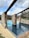 A rectangular swimming pool features two sections with the closer section having darker tiles and a more shallow appearance. The pool is bordered by stone and brick columns supporting wooden beams, creating an open, rustic look. Surrounding the pool is a patio area with lounge chairs, and the scene is set against a backdrop of cloudy sky and trees.