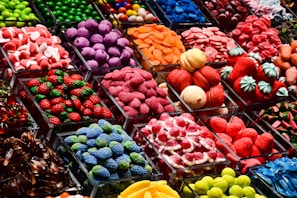 An assortment of colorful candy displayed in clear containers, featuring a variety of shapes and textures. The selection includes sweets that resemble berries, citrus slices, and marshmallows, arranged in an eye-catching, organized layout.