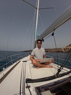A person with short dark hair and wearing sunglasses is sitting cross-legged on the deck of a sailboat, holding a laptop. The water surrounding the boat is calm, and rocky terrain is visible in the background. The person is wearing a striped T-shirt.