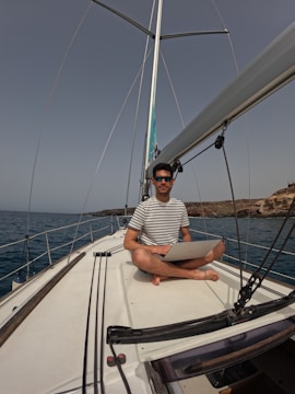 A person with short dark hair and wearing sunglasses is sitting cross-legged on the deck of a sailboat, holding a laptop. The water surrounding the boat is calm, and rocky terrain is visible in the background. The person is wearing a striped T-shirt.