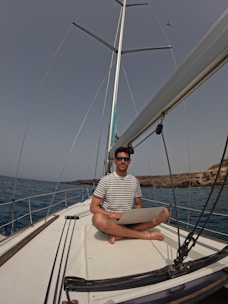 A man is sitting cross-legged on the deck of a sailboat, holding a laptop. He is wearing a striped shirt and sunglasses, and the surroundings include calm ocean waters and rocky coastal terrain. In the sky above, a small aircraft is visible.