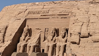 Several large, ancient statues carved into a sandstone cliff. These statues depict seated figures with detailed headdresses and attire, surrounded by intricate carvings and hieroglyphics. The setting is expansive and bathed in sunlight, highlighting the sandy texture and grandeur of the monument.