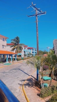 A street lined with palm trees and multi-story buildings featuring red-tiled roofs. A utility pole with power lines stretches across a bright blue sky, and paved walkways with plants in concrete planters are visible. The architecture appears tropical, and a small flag is seen in the distance.