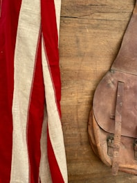 A vintage leather bag lies on a wooden surface, next to a fabric with red and white stripes.