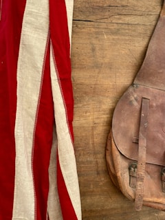A vintage leather bag lies on a wooden surface, next to a fabric with red and white stripes.