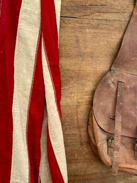 A vintage leather bag lies on a wooden surface, next to a fabric with red and white stripes.