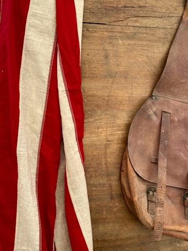 A vintage leather bag lies on a wooden surface, next to a fabric with red and white stripes.
