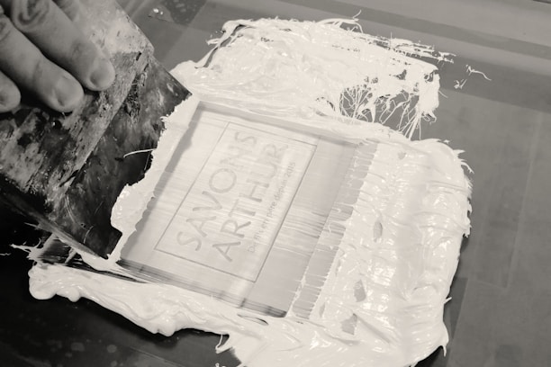 A hand is holding a squeegee, spreading white ink across a screen printing frame. The screen displays the text 'SAVON'S ARTHUR' and additional smaller writing is visible underneath. The ink is thickly applied and the process appears to be in progress, with some ink smeared beyond the frame.
