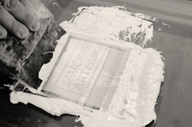 A hand is holding a squeegee, spreading white ink across a screen printing frame. The screen displays the text 'SAVON'S ARTHUR' and additional smaller writing is visible underneath. The ink is thickly applied and the process appears to be in progress, with some ink smeared beyond the frame.