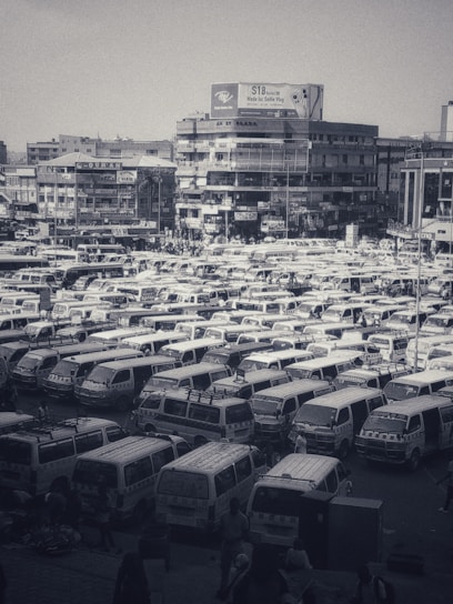 A densely packed parking lot filled with numerous white vans organized in rows. The background features multi-story buildings adorned with signs and billboards. The scene appears busy and urban, with clusters of people visible among the vehicles.