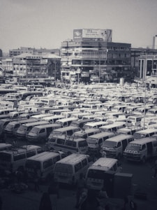 A densely packed parking lot filled with numerous white vans organized in rows. The background features multi-story buildings adorned with signs and billboards. The scene appears busy and urban, with clusters of people visible among the vehicles.