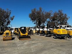 A collection of construction and industrial machinery is arranged outdoors on a paved surface, with a clear blue sky overhead. Various types of machines, including compactors, mini loaders, and portable generators, are lined up. Tall trees are visible in the background, providing a natural backdrop.