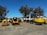 A collection of construction and industrial machinery is arranged outdoors on a paved surface, with a clear blue sky overhead. Various types of machines, including compactors, mini loaders, and portable generators, are lined up. Tall trees are visible in the background, providing a natural backdrop.