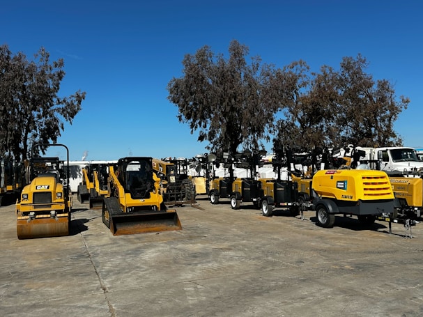 A collection of construction and industrial machinery is arranged outdoors on a paved surface, with a clear blue sky overhead. Various types of machines, including compactors, mini loaders, and portable generators, are lined up. Tall trees are visible in the background, providing a natural backdrop.