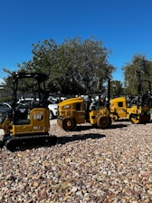 A row of yellow construction machinery is parked on a bed of rocks. The machines are industrial vehicles, likely used for road construction, and are aligned outdoors under a clear blue sky. Trees and a few parked cars are visible in the background.