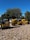 A row of yellow construction machinery is parked on a bed of rocks. The machines are industrial vehicles, likely used for road construction, and are aligned outdoors under a clear blue sky. Trees and a few parked cars are visible in the background.