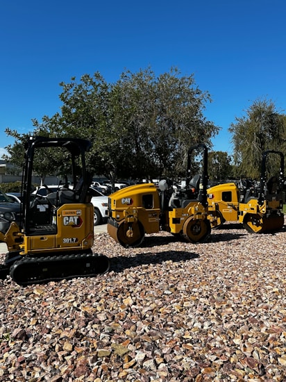 A row of yellow construction machinery is parked on a bed of rocks. The machines are industrial vehicles, likely used for road construction, and are aligned outdoors under a clear blue sky. Trees and a few parked cars are visible in the background.