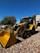 A yellow backhoe loader is parked on a gravel surface under a clear blue sky. The machinery is equipped with large tires and a front bucket. In the background, more construction equipment and a building with a textured facade are visible. Trees with green foliage provide additional background scenery.