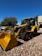 A yellow backhoe loader is parked on a gravel surface under a clear blue sky. The machinery is equipped with large tires and a front bucket. In the background, more construction equipment and a building with a textured facade are visible. Trees with green foliage provide additional background scenery.