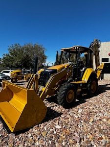 A yellow backhoe loader is parked on a gravel surface under a clear blue sky. The machinery is equipped with large tires and a front bucket. In the background, more construction equipment and a building with a textured facade are visible. Trees with green foliage provide additional background scenery.
