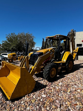 A yellow backhoe loader is parked on a gravel surface under a clear blue sky. The machinery is equipped with large tires and a front bucket. In the background, more construction equipment and a building with a textured facade are visible. Trees with green foliage provide additional background scenery.
