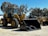 A large yellow construction vehicle with massive tires and a front loader attachment is parked on a paved surface. Several similar machinery units are aligned in the background under a clear blue sky. Tall trees with dense foliage surround the area.